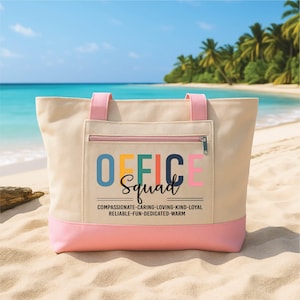 May include: A beige canvas tote bag with pink trim and handles, featuring the text "OFFICE Squad" in various colours. The bag has a zipped pocket. The text below reads "COMPASSIONATE-CARING-LOVING-KIND-LOYAL RELIABLE-FUN-DEDICATED-WARM". The bag is on a sandy beach.