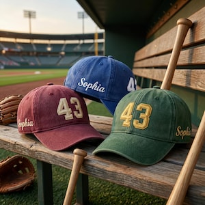 May include: Three baseball caps in maroon, blue, and green, each featuring the number 43 in a bold font on the front and the name Sophia embroidered in script on the side. The caps are displayed alongside wooden baseball bats and gloves.