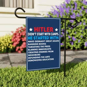 May include: A dark blue garden flag with white text and a white border. The text reads "HITLER DIDN'T START WITH CAMPS. HE STARTED WITH:" followed by a list of actions. The flag is displayed on a black metal stand in a garden setting.