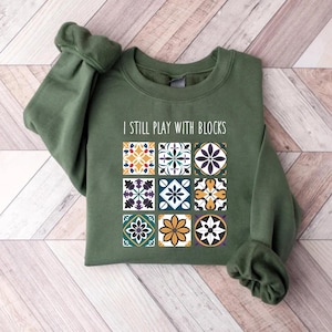 May include: A green sweatshirt with the text "I still play with blocks" and a pattern of colorful tiles.