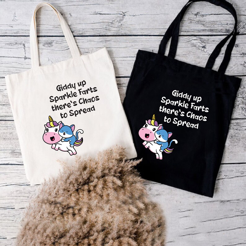 May include: Two tote bags with a unicorn and cat design. The text on the bags reads "Giddy up Sparkle Farts there's Chaos to Spread". One bag is white and the other is black.