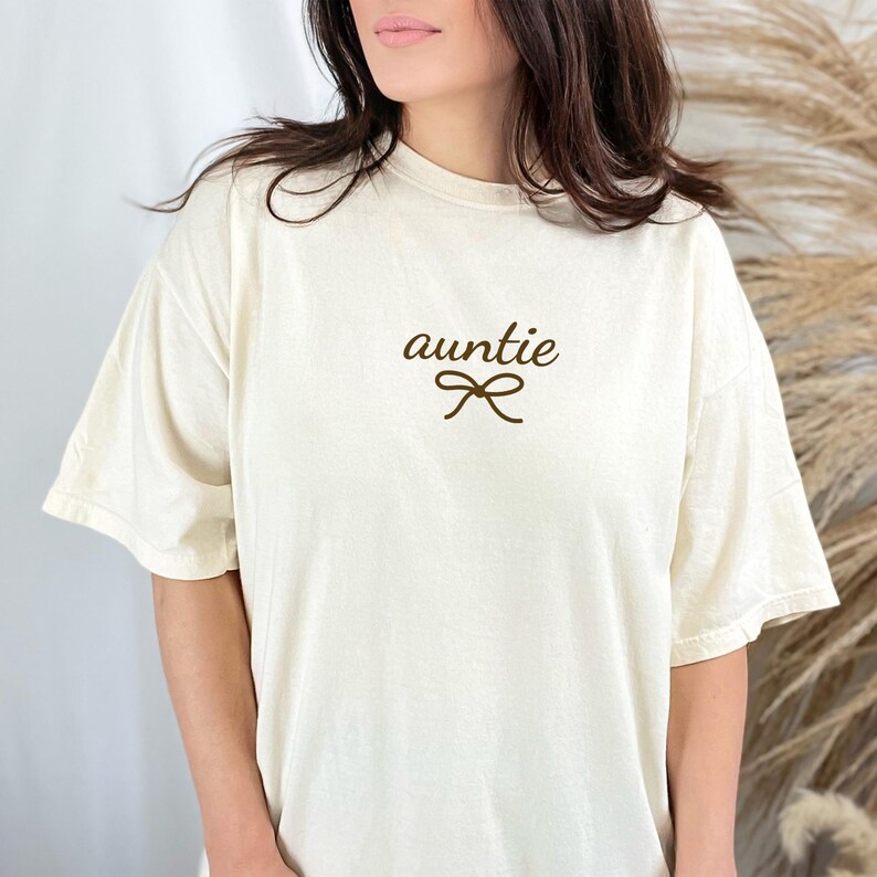 May include: Cream-colored t-shirt with the word "auntie" in a brown script font, accompanied by a bow graphic. The shirt has short sleeves and a relaxed fit, suitable for casual wear.