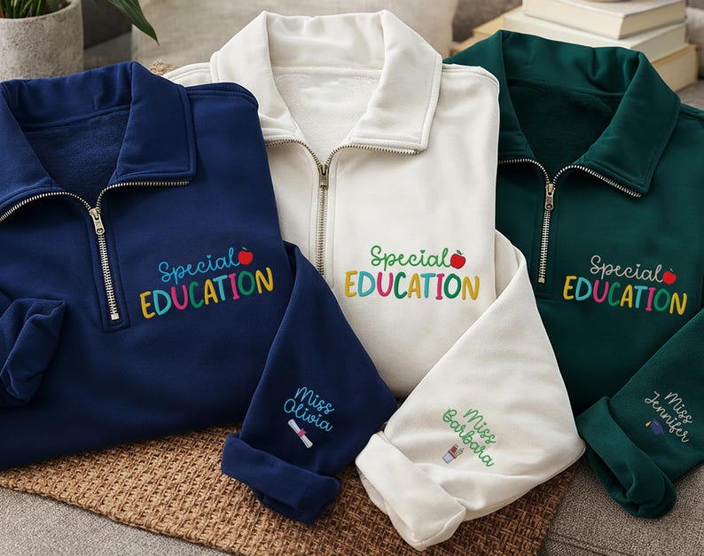 May include: Three quarter-zip sweatshirts in navy blue, white, and dark green. Each sweatshirt features the embroidered text "Special Education" with a red apple and colorful letters. The sleeves are rolled up, displaying names like "Miss Olivia," "Miss Barbara," and "Miss Jennifer."