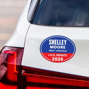May include: An oval car sticker with the text "SHELLEY MOORE WEST VIRGINIA U.S. SENATE 2026" in white, blue, and red. The sticker is attached to the rear of a white car, near the taillight.