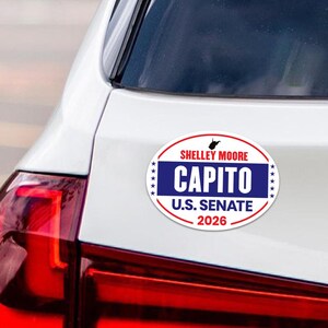 May include: An oval bumper sticker with the text "SHELLEY MOORE CAPITO U.S. SENATE 2026" in red, white, and blue. The sticker is attached to the rear of a white car, with a red taillight visible.
