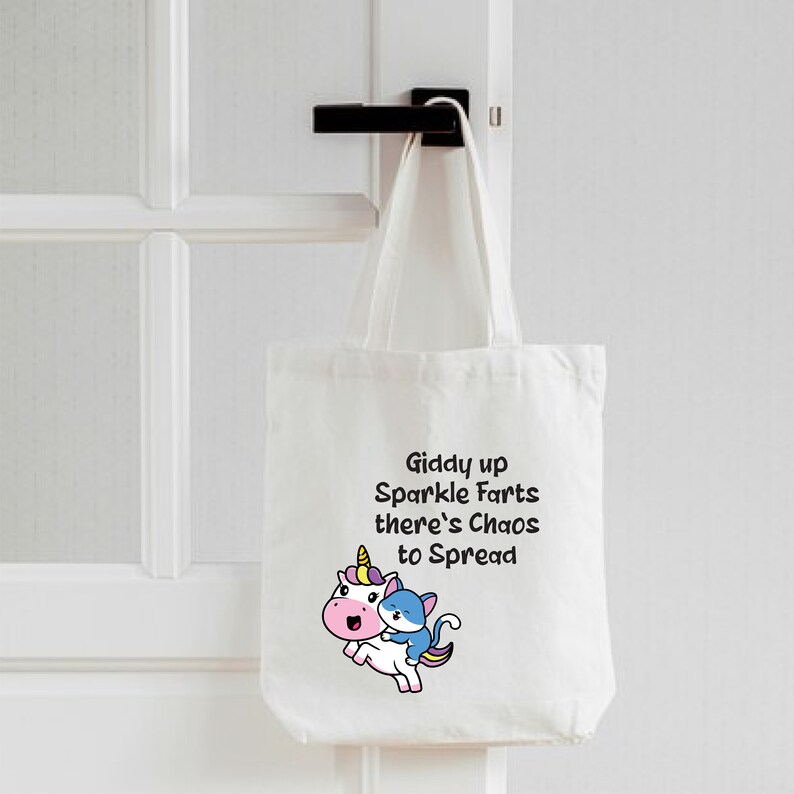 May include: White tote bag with a unicorn and a cat riding on its back. The bag has the text "Giddy up Sparkle Farts there's Chaos to Spread" printed on it.