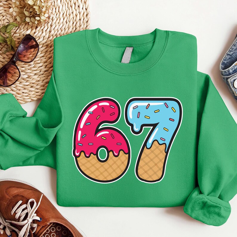 May include: A green sweatshirt features a colorful graphic of the number "67" designed to look like donuts and ice cream. The number "6" is pink with red sprinkles, and the number "7" is blue with sprinkles. The sweatshirt is made of a soft material.