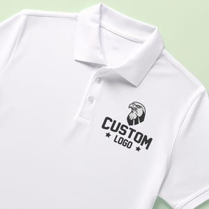 May include: White polo shirt with a collar and three buttons. The shirt features a black and white eagle logo and the words "CUSTOM LOGO" printed below it. The shirt is made of a smooth, likely synthetic fabric.