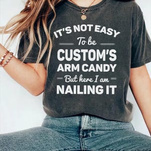 May include: Dark gray t-shirt with white text that reads "IT'S NOT EASY To be CUSTOM'S ARM CANDY - But here I am - NAILING IT". The shirt is a casual style.