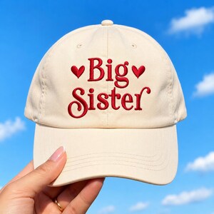 May include: A cream-colored baseball cap featuring the embroidered red text Big Sister centered on the front panel. The text is flanked by two small red embroidered hearts, one on each side. The cap has a structured crown and a curved brim.