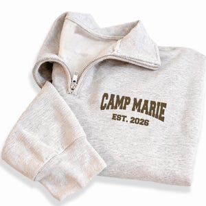 Peut inclure: Un sweat-shirt gris clair avec un col zippé et des poignets côtelés. Le texte "CAMP MARIE EST. 2026" est imprimé en marron sur le devant. Le sweat-shirt est fait d'une matière douce et confortable.