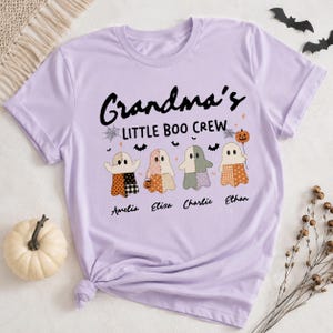 May include: A light purple short-sleeve t-shirt featuring the black text Grandma's LITTLE BOO CREW. Below the text are four ghost illustrations with patterned bodies, small bats, a spiderweb, and a pumpkin balloon. The names Amelia, Elisa, Charlie, and Ethan are printed in black script below each ghost.