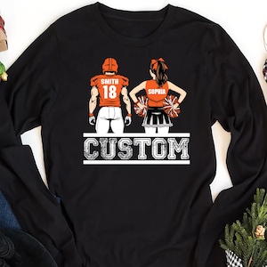 May include: Black long-sleeve shirt featuring a graphic of a football player and a cheerleader. The football player's jersey has the name "SMITH" and the number "18". The cheerleader's top has the name "SOPHIA". The word "CUSTOM" is printed below the figures.
