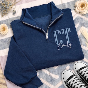 May include: Navy blue quarter-zip sweatshirt with the initials "CT" and the name "Emily" in light blue. The sweatshirt has a silver zipper and is paired with black and white sneakers.