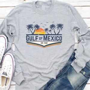 May include: Gray long-sleeve t-shirt with a vintage-style graphic of palm trees, a sunset, and the text "GULF of MEXICO EST. 1550".