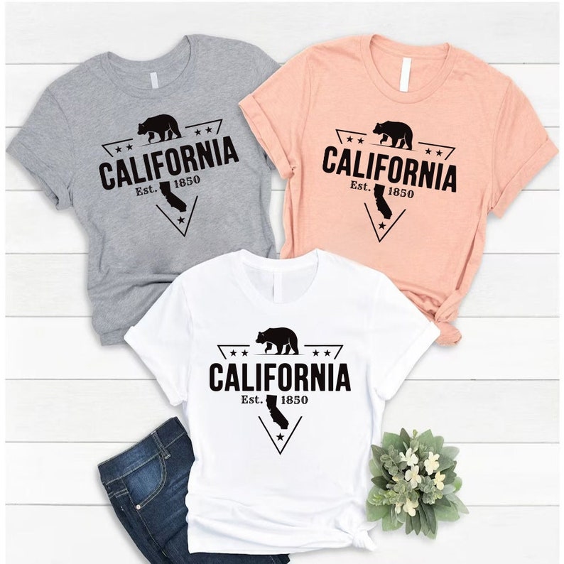 May include: Three t-shirts with a black and white graphic design of a bear and the text "CALIFORNIA Est. 1850". The t-shirts are in grey, peach, and white.