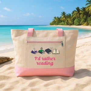 May include: A beige canvas tote bag with pink trim and handles, featuring a front zippered pocket. The design includes illustrations of stacked books, a coffee mug, glasses, and a plant, with the pink text "I'd rather reading" printed below.