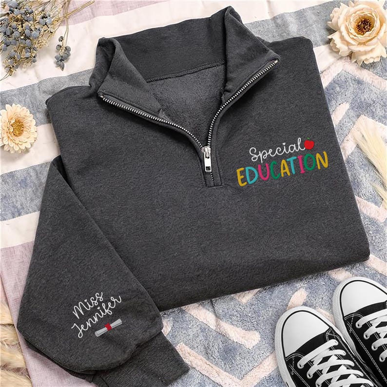 May include: Dark gray quarter-zip sweatshirt with the words "Special Education" embroidered in colorful letters. The sleeve features the name "Miss Jennifer" and a small graduation scroll. Black and white sneakers are at the bottom.