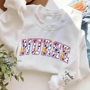 May include: White sweatshirt with a floral print design that spells out "NURSE" in a purple outline. The sweatshirt has a small embroidered stethoscope and heart on the sleeve with the word "Amelia" written next to it.