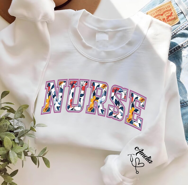 May include: White sweatshirt with a floral print design that spells out "NURSE" in a purple outline. The sweatshirt has a small embroidered stethoscope and heart on the sleeve with the word "Amelia" written next to it.