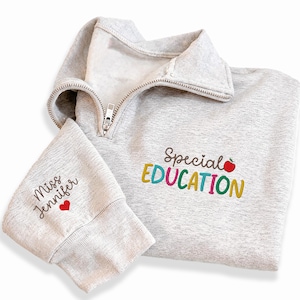 May include: A light gray quarter-zip sweatshirt with the words "Special Education" embroidered in colorful letters and a red apple. The left sleeve has "Miss Jennifer" embroidered with a red heart. The sweatshirt has a silver zipper.