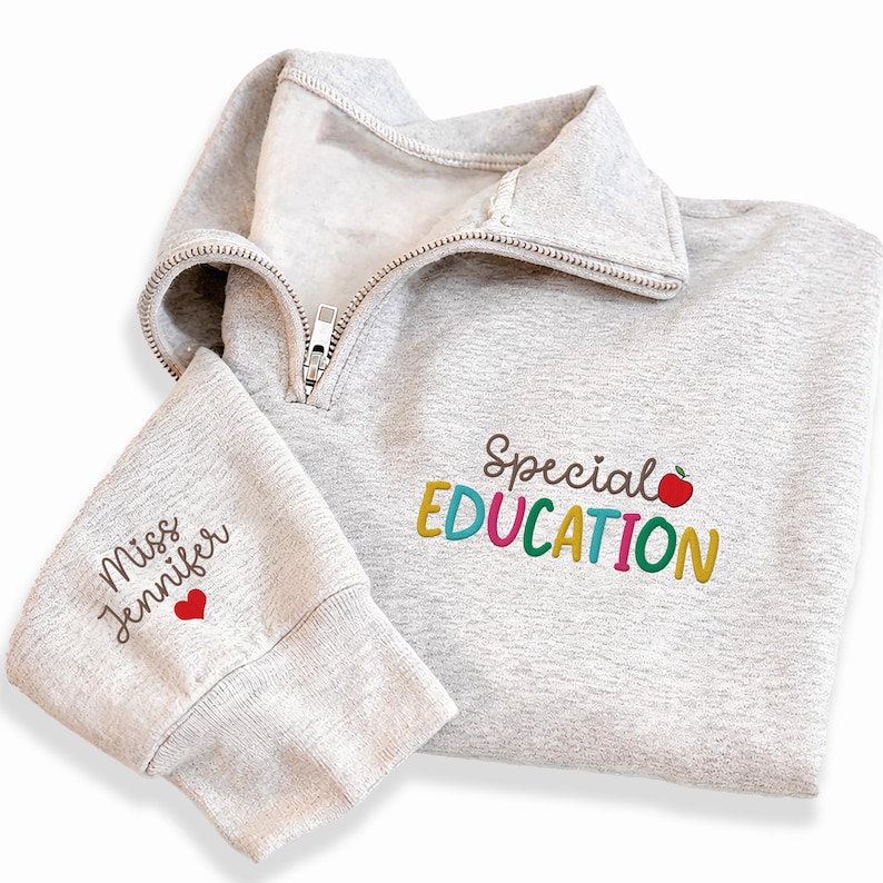 May include: A light gray quarter-zip sweatshirt with the words "Special Education" embroidered in colorful letters and a red apple. The left sleeve has "Miss Jennifer" embroidered with a red heart. The sweatshirt has a silver zipper.