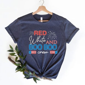May include: Navy blue t-shirt with the text "Red White and Boo Boo crew" in red, white, and blue lettering. The shirt features a small American flag graphic on each side of the text and a firework graphic above the text.