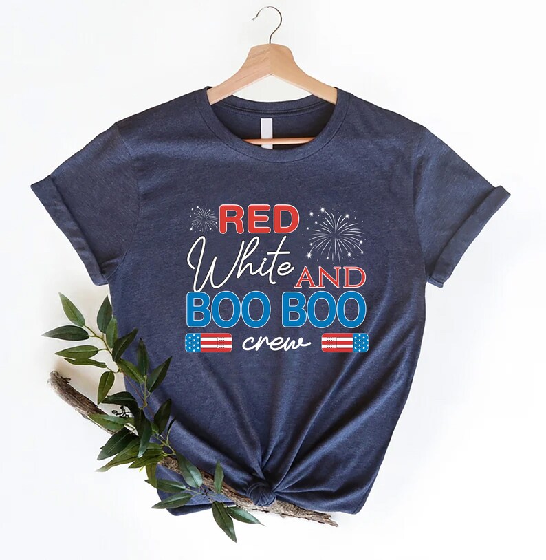 May include: Navy blue t-shirt with the text "Red White and Boo Boo crew" in red, white, and blue lettering. The shirt features a small American flag graphic on each side of the text and a firework graphic above the text.