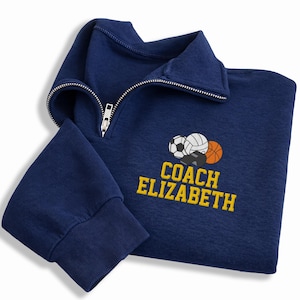 May include: Navy blue quarter-zip sweatshirt with the text "COACH ELIZABETH" in yellow. The sweatshirt features embroidered sports balls and a whistle design. The sleeve cuff is ribbed.