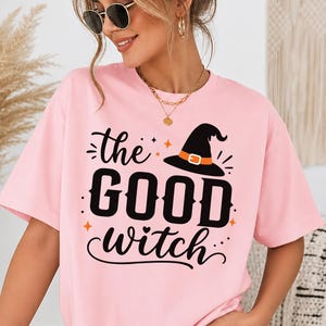 May include: A light pink short-sleeved t-shirt featuring a black graphic print that reads the GOOD witch in a mix of bold and cursive fonts. The design includes a black witch hat with an orange band and small orange star accents.