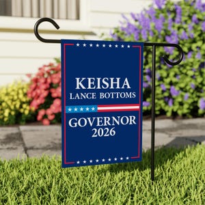 May include: A blue garden flag with the text "KEISHA LANCE BOTTOMS GOVERNOR 2026" in white. The flag has a red border and a design of stars and stripes. The flag is displayed on a black metal stand in a grassy yard.