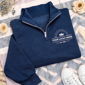 May include: A navy blue quarter-zip sweatshirt featuring a silver zipper and a white graphic print on the chest. The design includes a crown icon, sunburst lines, and the text "YOUR LOGO HERE" followed by "EST. 2026" in a clean, sans-serif font.