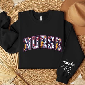 May include: Black sweatshirt with a floral print "NURSE" design and the name "Amelia" with a stethoscope and heart on the sleeve.