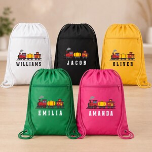 May include: Five drawstring backpacks in white, black, yellow, green, and pink, each featuring a front zipper pocket and a graphic of a red train with a yellow tank car. The names Williams, Jacob, Oliver, Emilia, and Amanda are printed below.