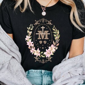 Op de afbeelding: Zwart T-shirt met een bloemenmotief en de tekst "O Maria, ontvangen zonder zonde, bid voor ons die onze toevlucht tot u nemen." Het ontwerp bevat een gestileerde "M" met een hart en doornen.