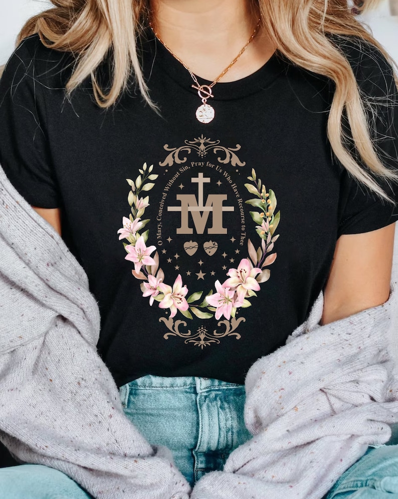 Op de afbeelding: Zwart T-shirt met een bloemenmotief en de tekst "O Maria, ontvangen zonder zonde, bid voor ons die onze toevlucht tot u nemen." Het ontwerp bevat een gestileerde "M" met een hart en doornen.