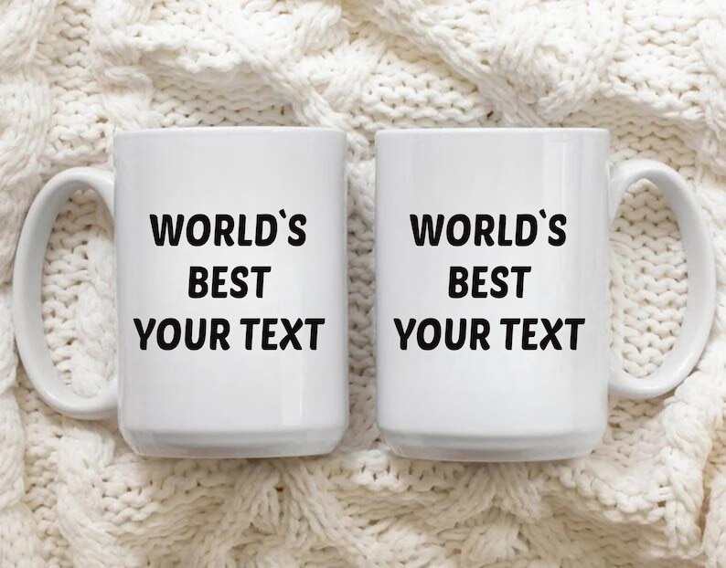 May include: Two white ceramic mugs with black text that reads "World's Best Your Text". The mugs are on a white knitted surface.