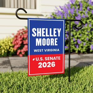 May include: A garden flag with the text "SHELLEY MOORE WEST VIRGINIA U.S. SENATE 2026" in white letters on a blue and red background. The flag is displayed on a black metal stand in a grassy garden.