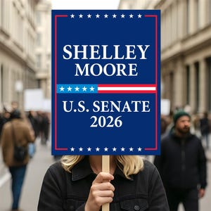 May include: A blue sign with the name "SHELLEY MOORE" in white capital letters, and "U.S. SENATE 2026" below. The sign has a red border and a design of stars and stripes. Held by a person in a crowd.