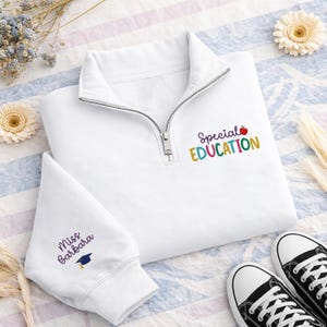 May include: White quarter-zip sweatshirt with the words "Special EDUCATION" in colorful letters and an apple graphic. The sleeve has the text "Miss Barbara" and a graduation cap. Black and white sneakers are at the bottom.