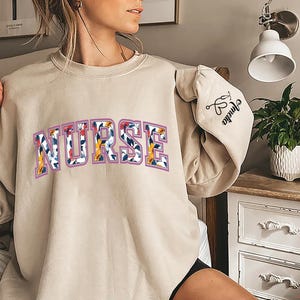 May include: A beige sweatshirt with the word "NURSE" printed in colorful letters. The sweatshirt has a floral pattern and the words "Grandma" and a heart symbol on the sleeve.