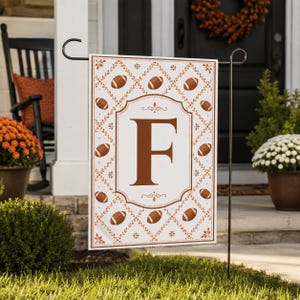 May include: A rectangular garden flag featuring a white background with a repeating pattern of brown footballs and leafy vines. A large, centred brown letter F is enclosed within a decorative border. The flag hangs from a black metal stand.