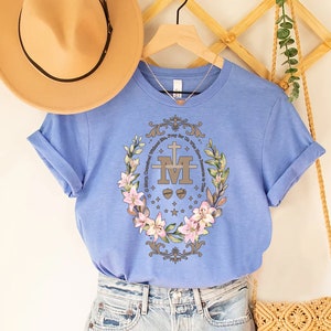 Op de afbeelding: Een lichtblauw T-shirt met een bloemenmotief en de tekst "Maria, ontvangen zonder zonde, bid voor ons die onze toevlucht tot u nemen." Het ontwerp bevat een gestileerde "M" met een kruis en twee harten.