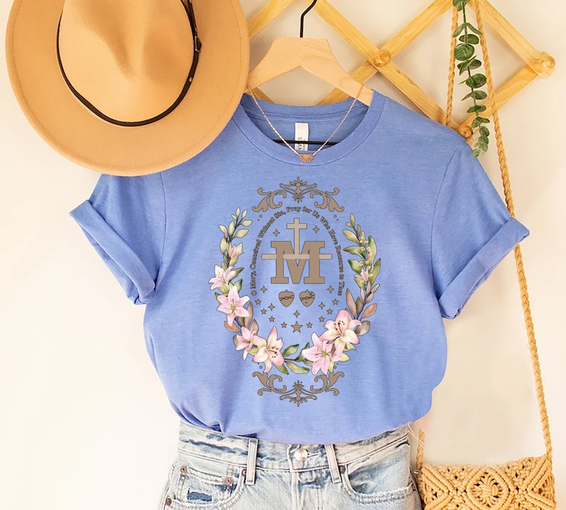 Op de afbeelding: Een lichtblauw T-shirt met een bloemenmotief en de tekst "Maria, ontvangen zonder zonde, bid voor ons die onze toevlucht tot u nemen." Het ontwerp bevat een gestileerde "M" met een kruis en twee harten.