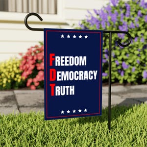 May include: A navy blue garden flag with the words "FREEDOM DEMOCRACY TRUTH" in white and red lettering. The flag is bordered in red and has white stars at the top and bottom. It is displayed on a black metal stand in a garden.