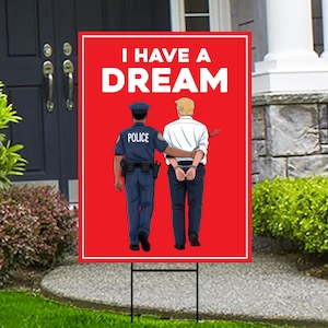 May include: A red yard sign with the words "I HAVE A DREAM" in white. The sign features an illustration of a police officer with a man in handcuffs. The officer has his arm around the man's shoulder.