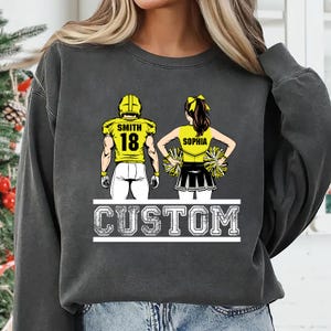 May include: Dark gray sweatshirt featuring a football player and cheerleader design. The football player wears a yellow jersey with the number 18 and the name "Smith". The cheerleader wears a yellow top with the name "Sophia". The word "CUSTOM" is printed below.