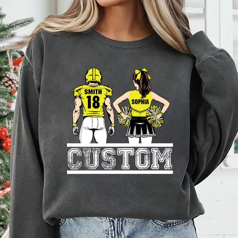 May include: Dark gray sweatshirt featuring a football player and cheerleader design. The football player wears a yellow jersey with the number 18 and the name "Smith". The cheerleader wears a yellow top with the name "Sophia". The word "CUSTOM" is printed below.