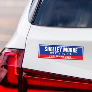 May include: A rectangular bumper sticker with the text "SHELLEY MOORE" in white letters on a blue background. Below, "WEST VIRGINIA" and "U.S. SENATE 2026" are displayed. The sticker has a red border and is affixed to a white car.
