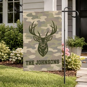 May include: A rectangular garden flag featuring a camouflage pattern in shades of green, brown, and tan. Centered on the flag is a dark green silhouette of a deer head with antlers. Below the deer, the text THE JOHNSONS is printed in dark green.